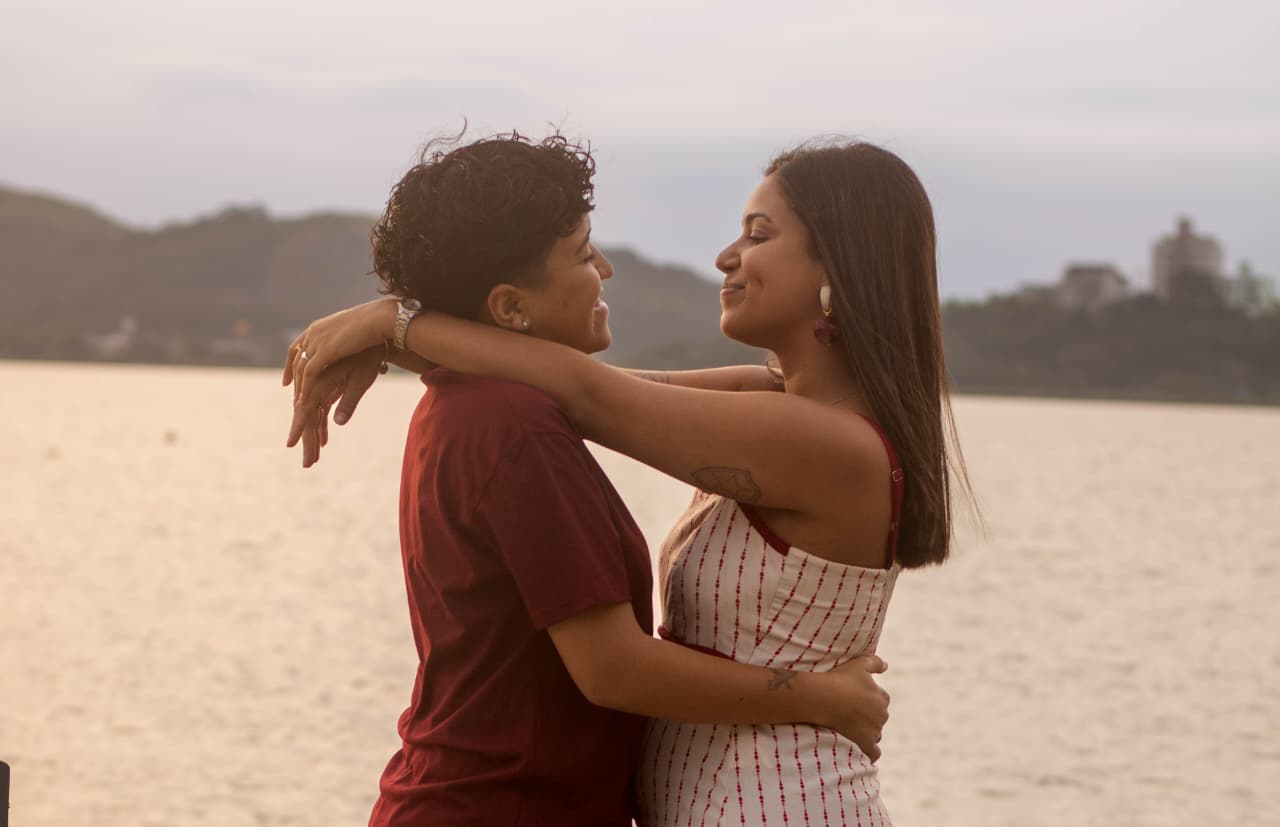 Casal abraçado de frente, sorrindo à beira-mar